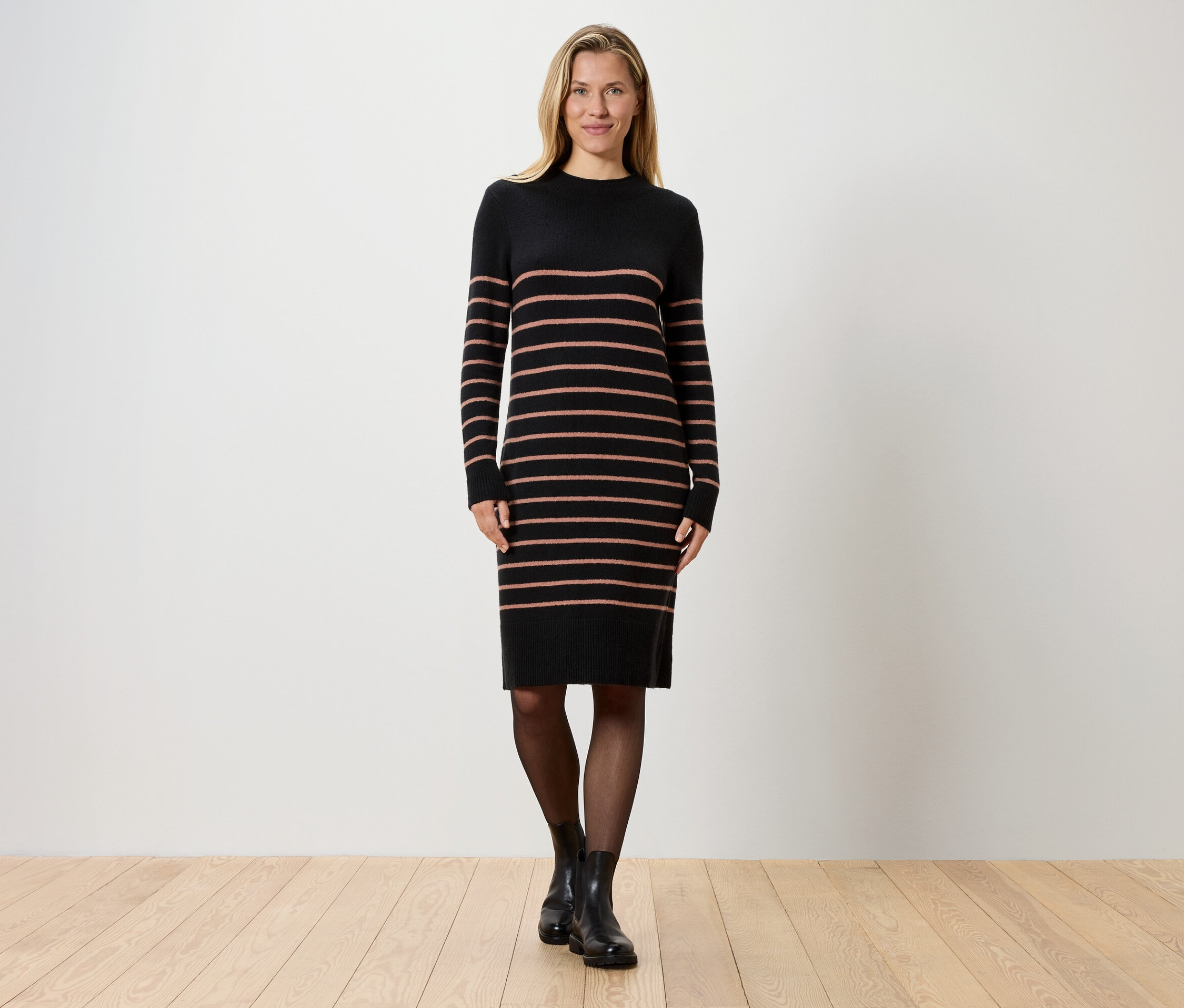 Blondínka stojí pred bielou stenou a má na sebe pruhované čierne pletené šaty, čierne pančuchy a čierne topánky.