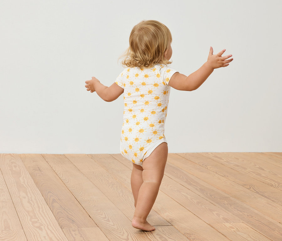 Malé dieťa s blond vlasmi v body s potlačou slnečníc stojí na drevenej podlahe.