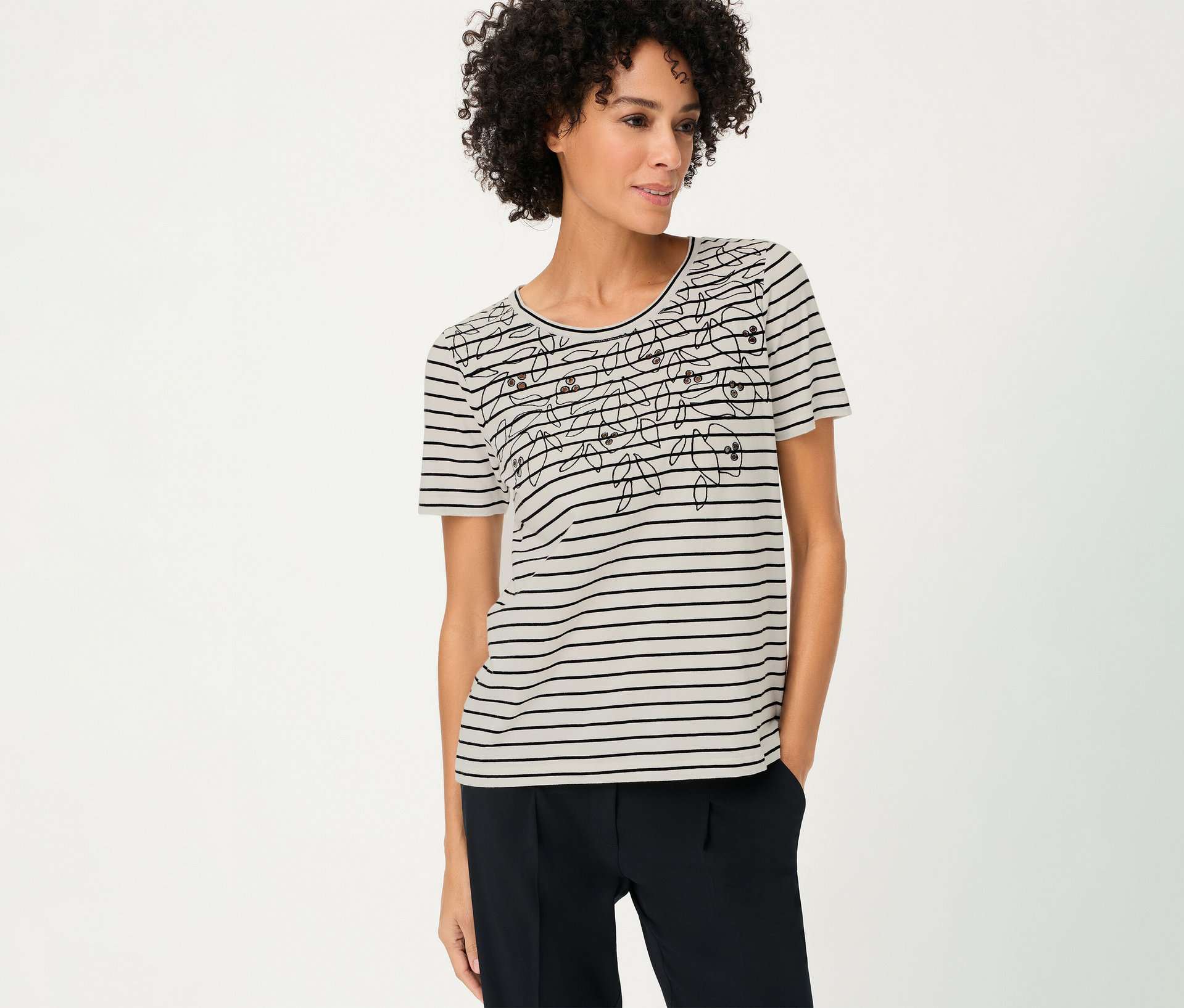 Žena s kučeravými vlasmi má na sebe pásikavé tričko Olsen s kvetinovým vzorom a tmavé nohavice.