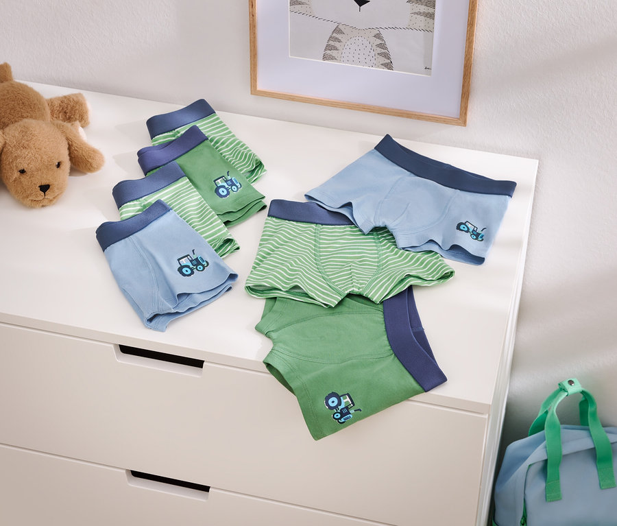 Fotografia svetlomodrých a zelených chlapčenských boxeriek s motívom traktora na komode, vedľa plyšového psa a modrej cestovnej tašky.