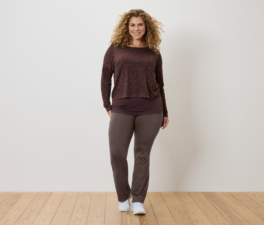 Žena s kučeravými vlasmi má na sebe hnedú vzorovanú blúzku s dlhým rukávom, tmavší top a hnedé nohavice a biele tenisky.