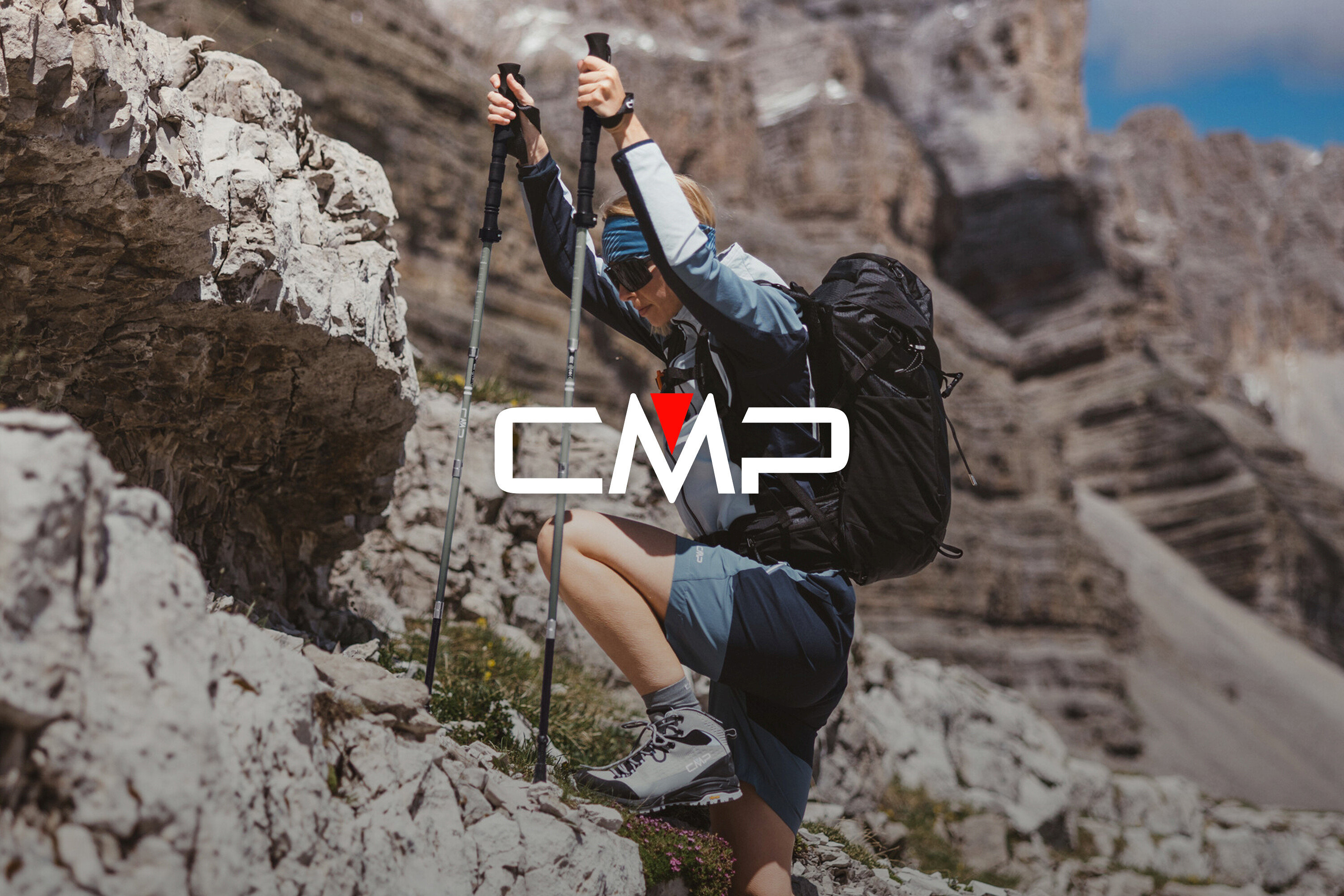 Žena s batohom a trekingovými palicami lezie po skalách. Má na sebe trekové topánky, šortky a bundu.