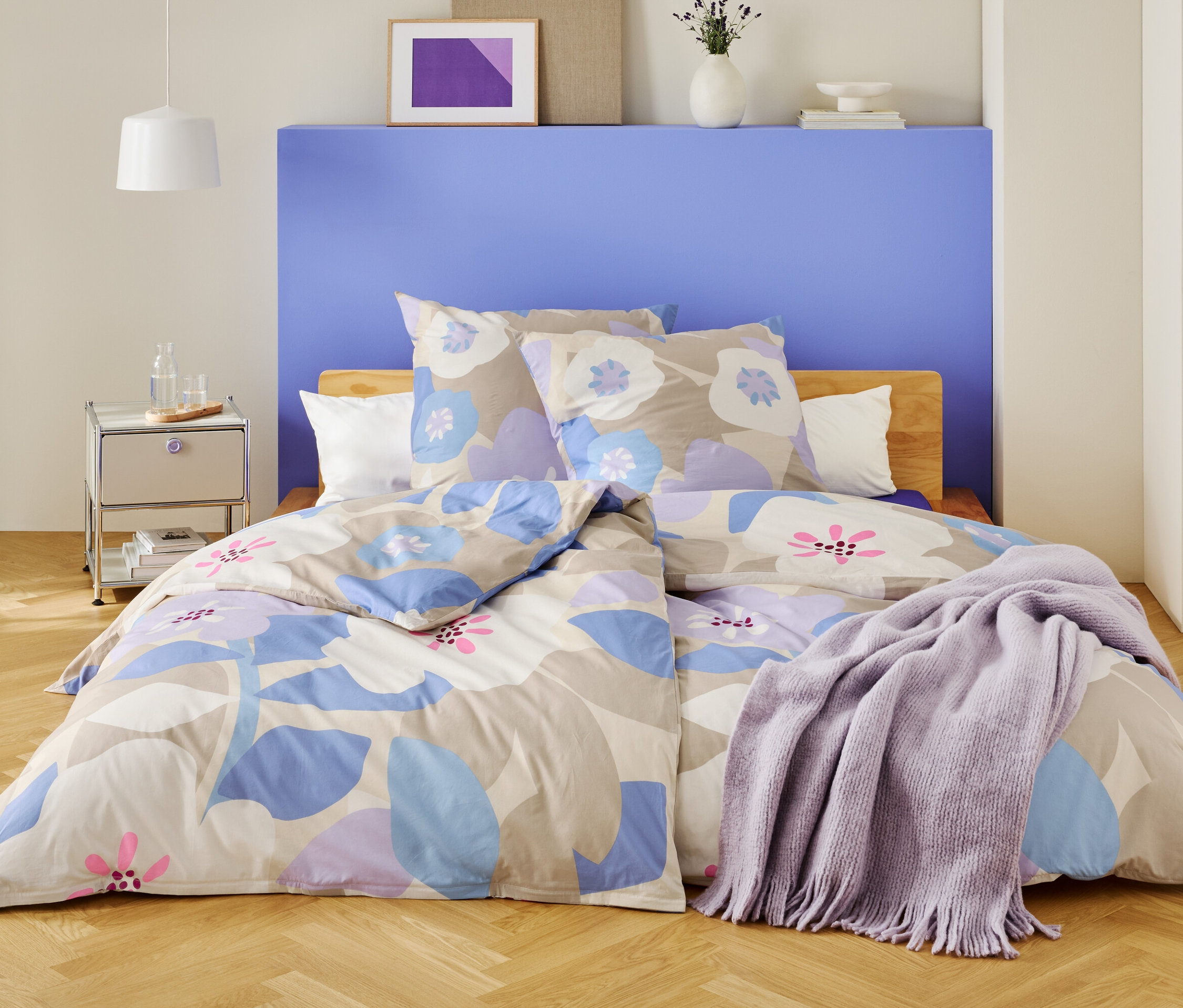 Spálňa s posteľou prikrytou kvetinovou posteľnou bielizňou a fialovou dekou.