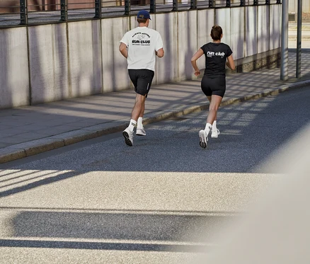 Muž v bielom športovom tričku s nápisom „RUN CLUB“, čiernych športových šortkách a ponožkách Coffee Crew a žena v čiernom športovom tričku s nápisom „RUN CLUB“ a krátkych športových legínsach ActivePower bežia po ceste.