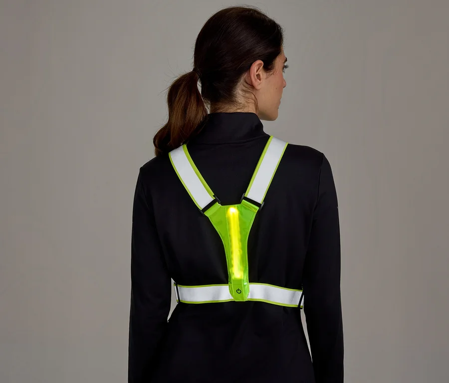Žena zozadu v čiernom tričku s dlhým rukávom a neónovo žltej reflexnej veste s vertikálnym svetelným pásom na chrbte.