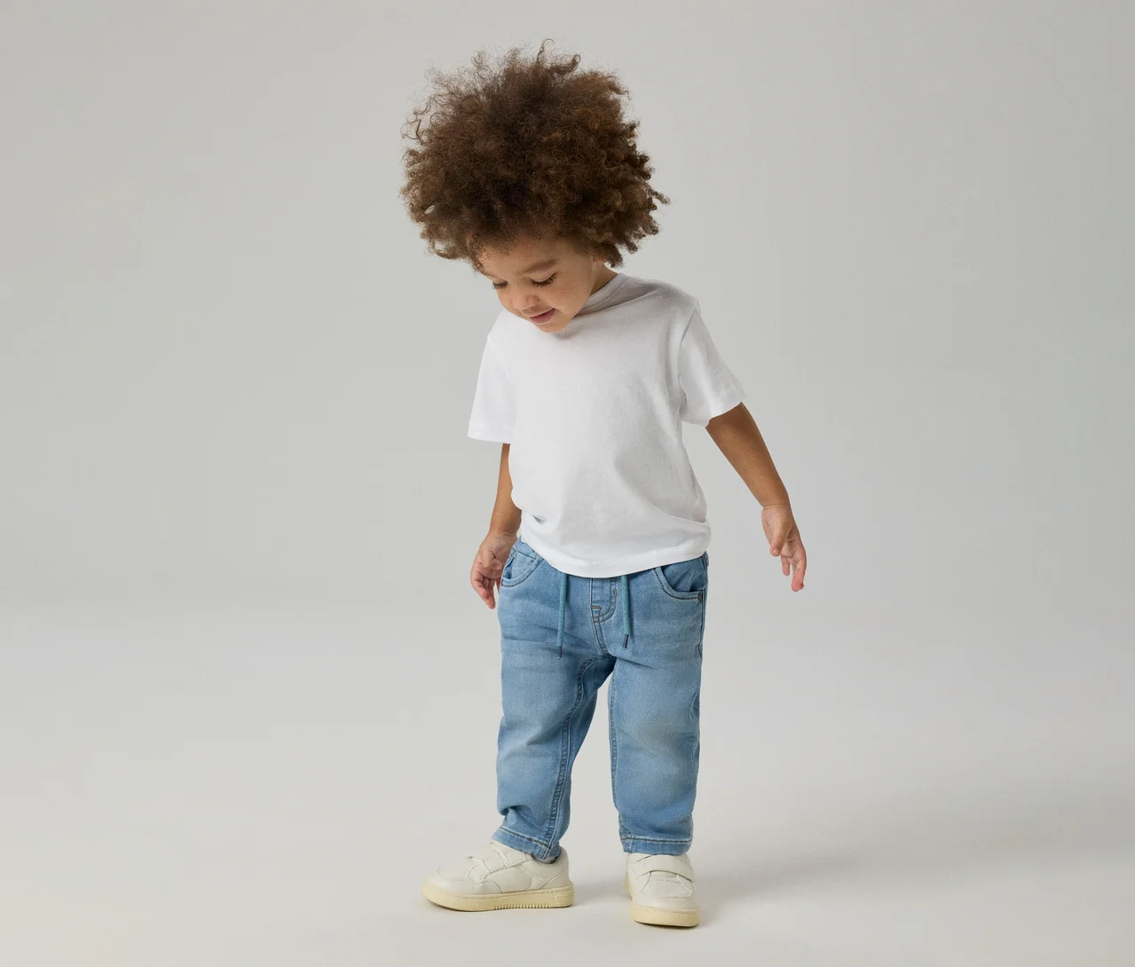 Malé dieťa s kučeravými vlasmi v bielom tričku, svetlomodrých NAME IT Slim Sweat Jeans a svetlých teniskách, pozerajúce sa dole.