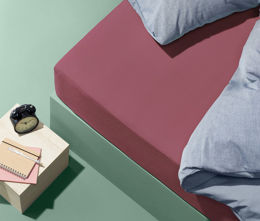 Ružová napínacia plachta z česanej bavlny štandardnej veľkosti na posteli s modrými vankúšmi a paplónom. Na nočnom stolíku leží budík a zápisník.