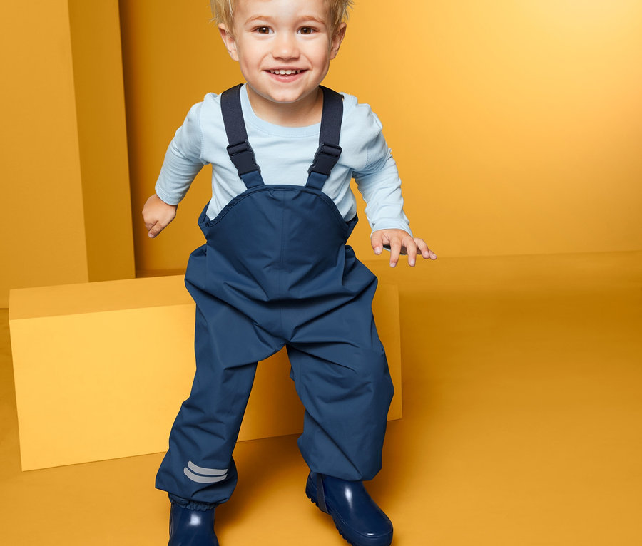 Usmievajúce sa batoľa stojí na žltom pozadí a má na sebe modré nohavice do dažďa.