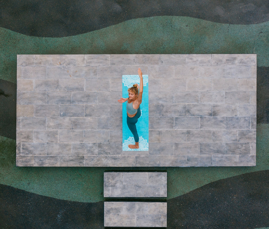 Pohľad zhora na ženu cvičiacu jogu na modrej podložke na šedej tehlovej plošine.