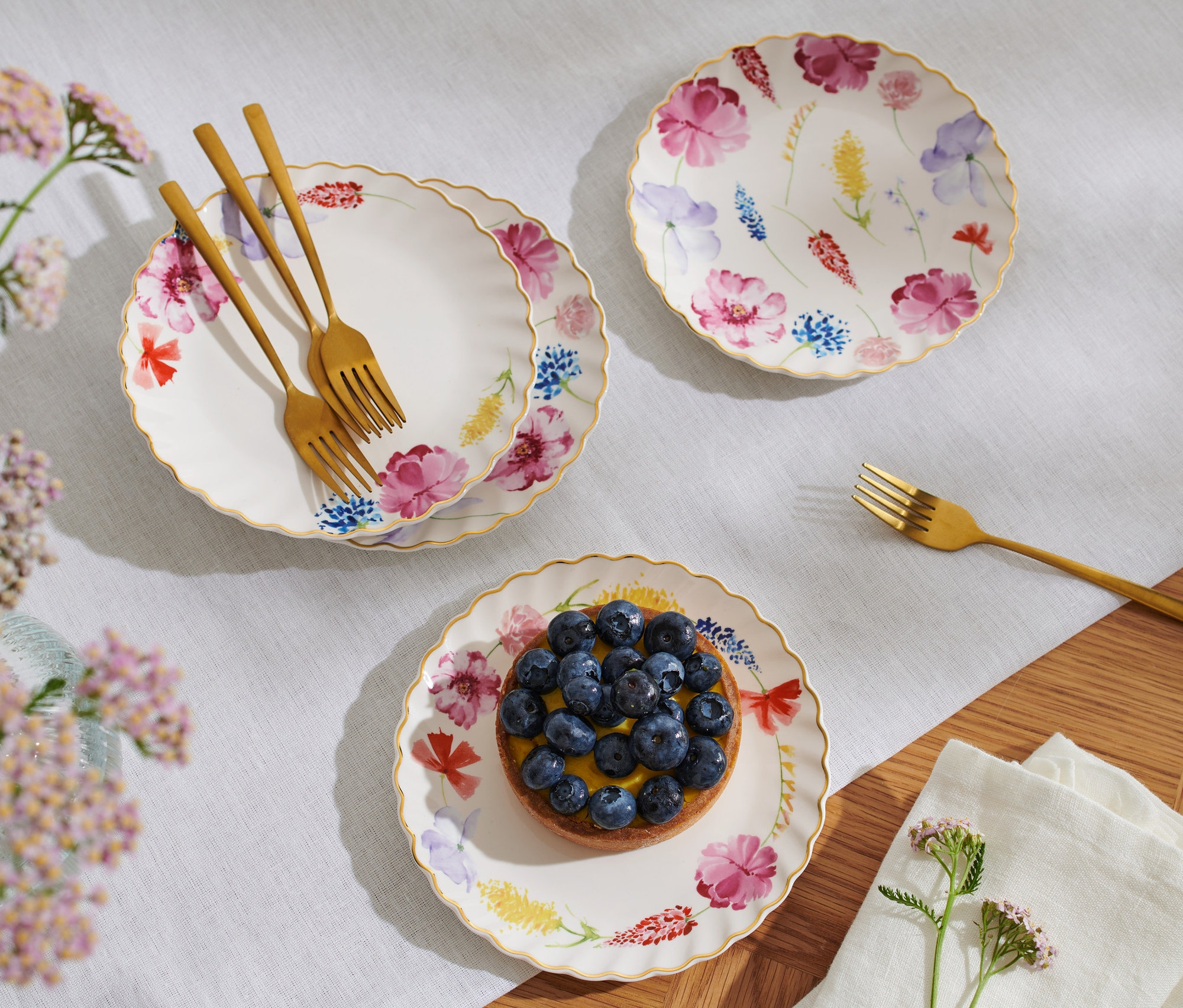 Tri dezertné taniere s lesklým zlatým dekorom a kvetinovým vzorom, na jednom je čučoriedkový koláč, vedľa ležia zlaté vidličky.