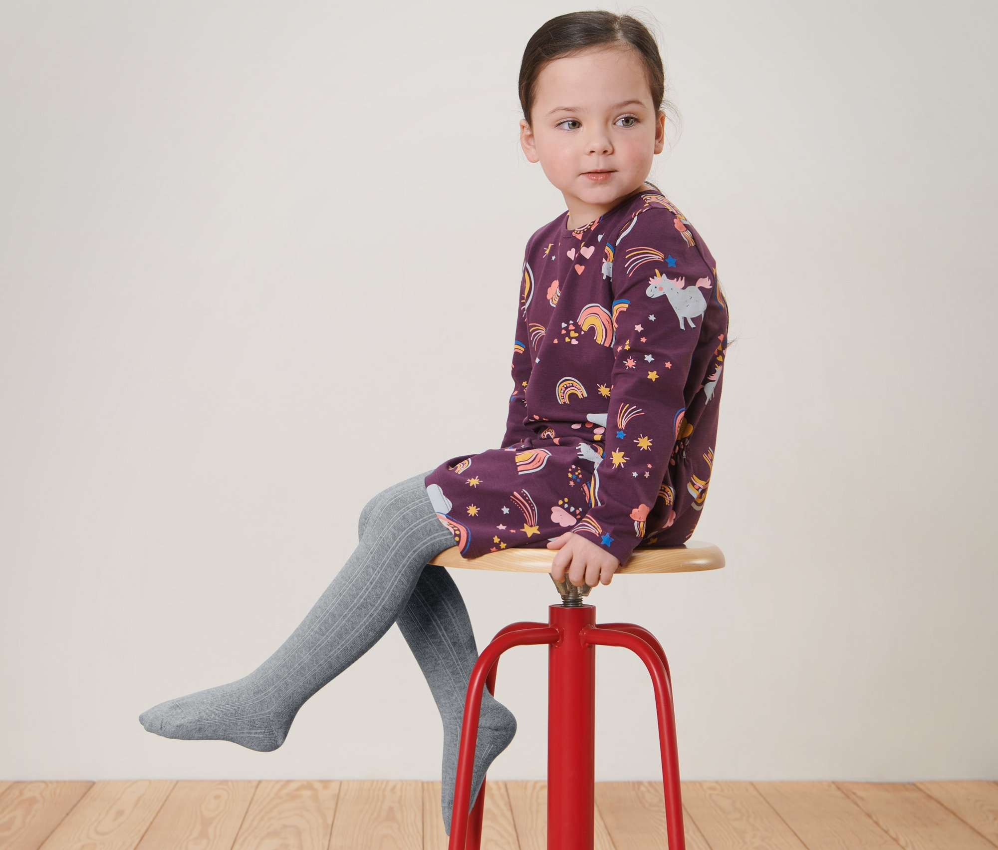 Dievča sedí na červenej stoličke a má na sebe šaty s potlačou a sivé detské pančuchové nohavice, 2 páry.