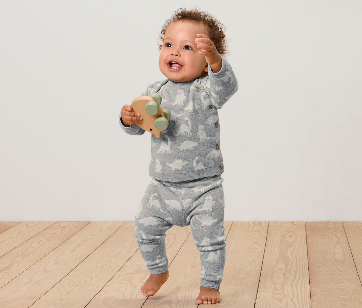 Dieťa stojí na drevenej podlahe a drží drevenú hračku. Má na sebe detskú pletenú súpravu.