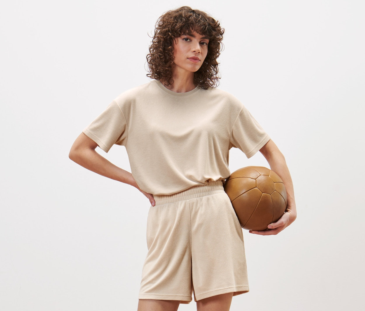 Žena pózuje v štúdiu a má na sebe športové froté šortky a drží hnedú futbalovú loptu.