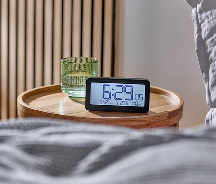 Digitálny LCD budík riadený rádiovým signálom na drevenom stolíku vedľa postele.