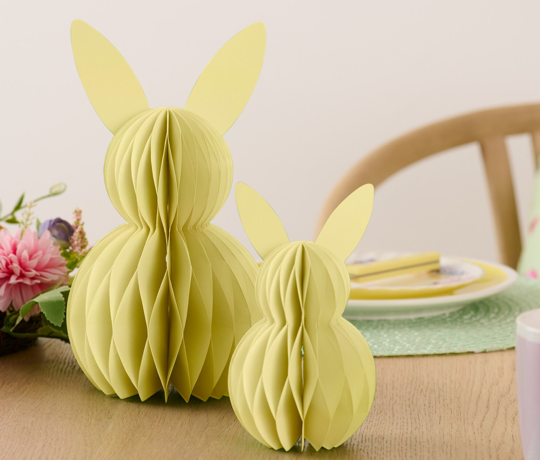 Dva papierové veľkonočné zajace stoja na stole vedľa kvetov a tanierov.