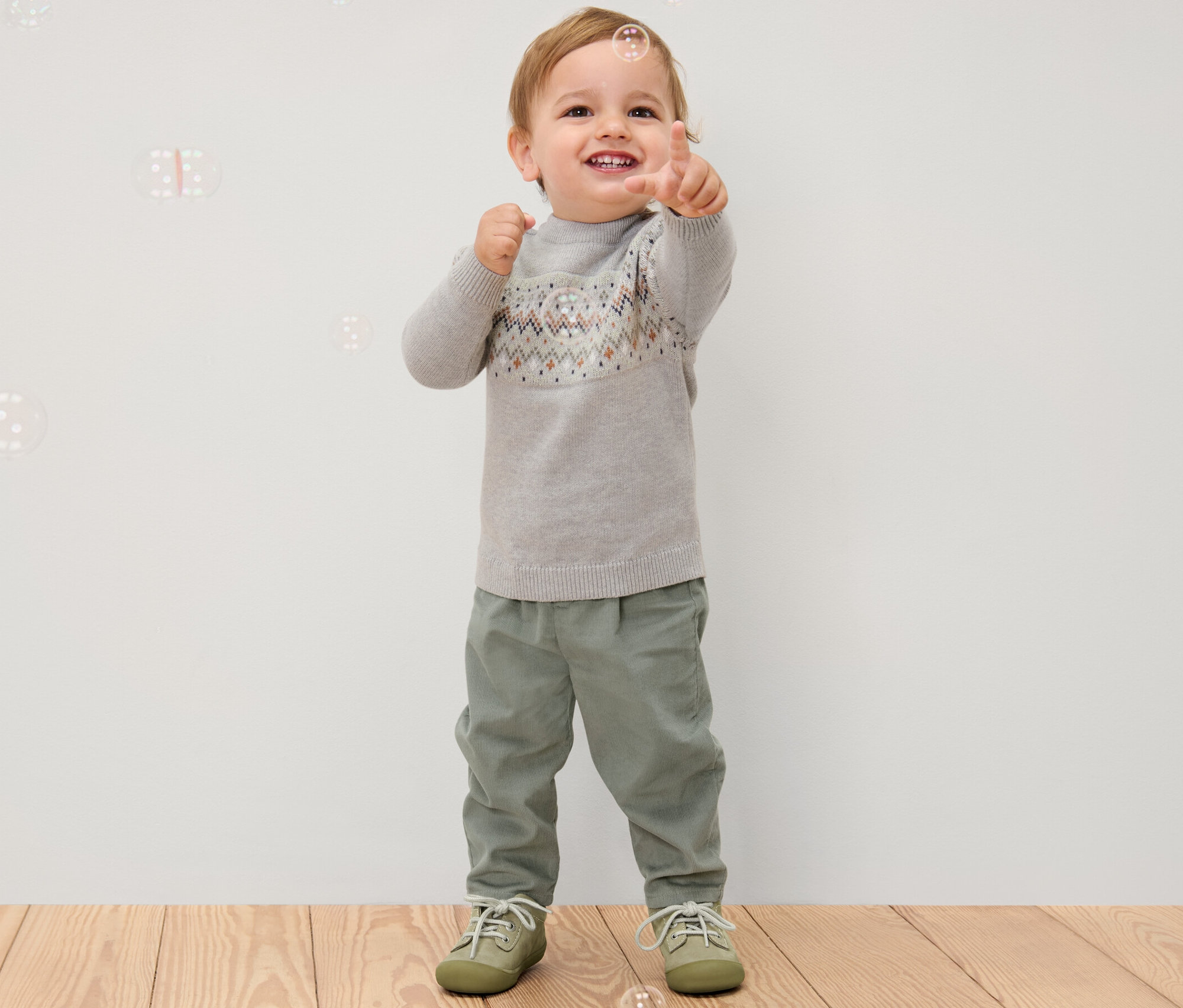 Chlapček ukazuje prstom, oblečený do sivého svetra a detských menčestrových pohodlných nohavíc.