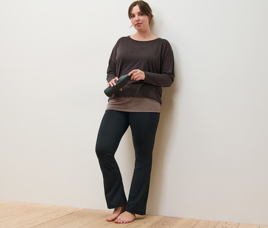 Žena opretá o stenu drží hnedé športové tričko 2 v 1 a sťahovacie športové nohavice.