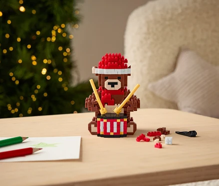 Kockový medveď s červenou čiapkou a šálom hrá na bubon paličkami na stole.