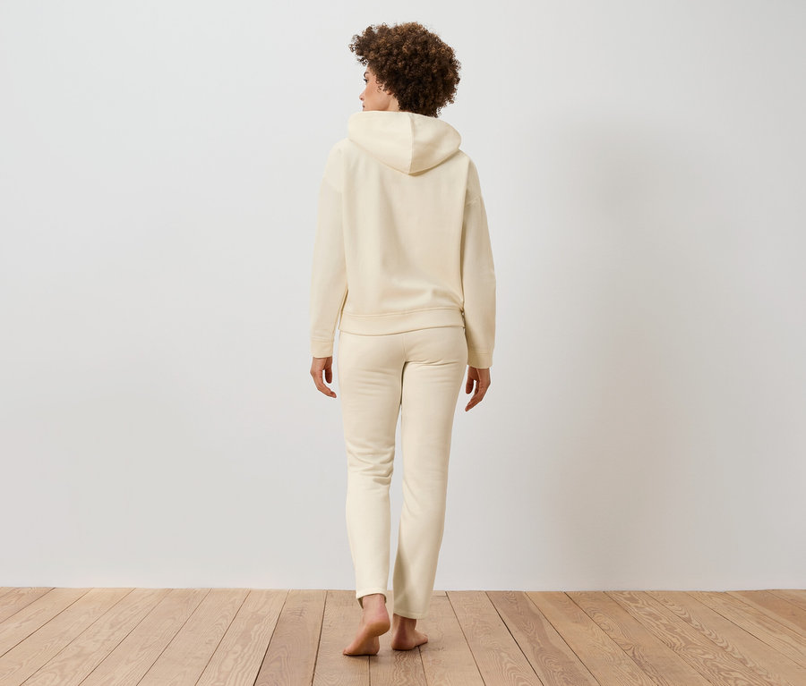 Žena s kučeravými vlasmi stojí chrbtom, oblečená do béžovej mikiny s kapucňou a teplákov na drevenej podlahe.