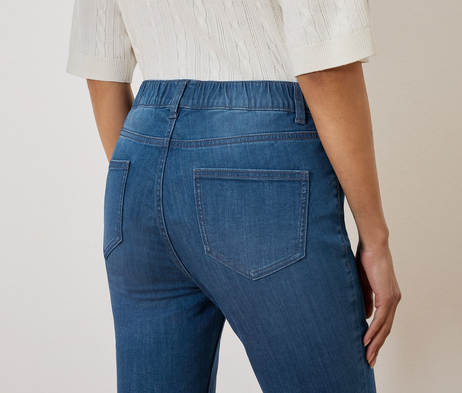 Detailný záber na chrbát ženy v modrých džínsoch s elastickým pásom a bielej pletenej blúzke.