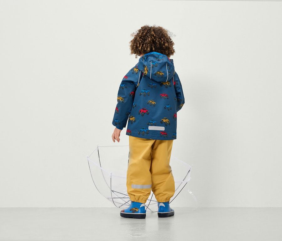 Chlapec v bunde s potlačou áut a žltých nohaviciach stojí chrbtom k fotoaparátu vedľa priehľadného dáždnika.