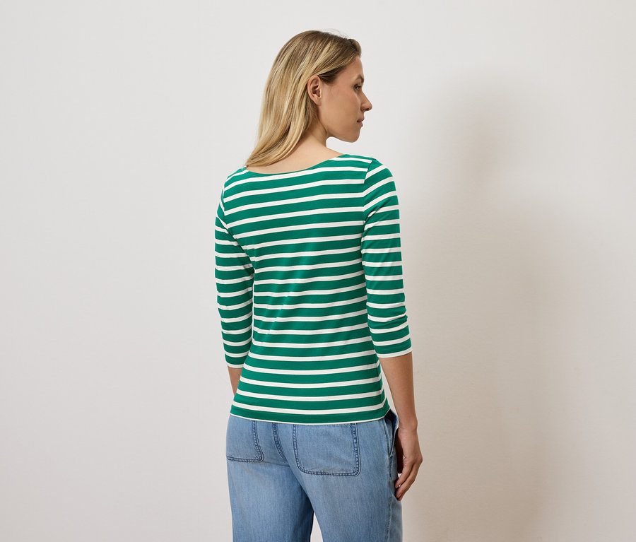 Žena s blond vlasmi stojí chrbtom k fotoaparátu a má na sebe tričko s trojštvrťovým rukávom so zelenými a bielymi pruhmi a modré džínsy.