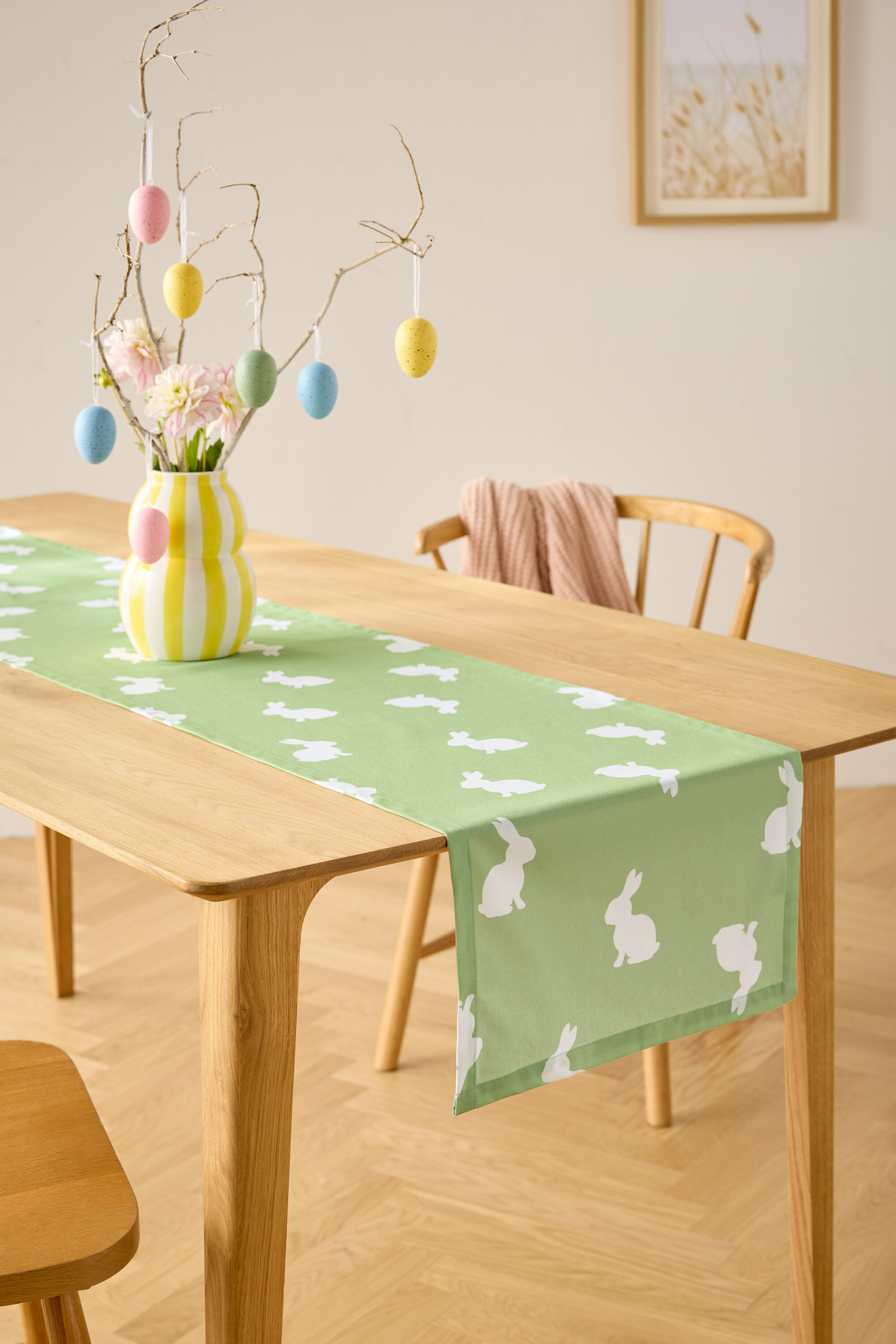 Jedálenský stôl so svetlozeleným behúňom s bielymi motívmi zajačikov, váza s kvetmi a dekorácia s veľkonočnými vajíčkami.
