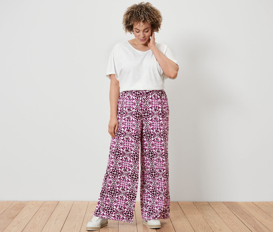 Žena v bielom tričku z ľanovej zmesi a nohaviciach v štýle palazzo s potlačou, bordovej, stojí na drevenej podlahe.