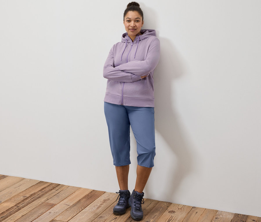 Žena stojí s prekríženými rukami a má na sebe fialovú mikinu a funkčné capri nohavice.