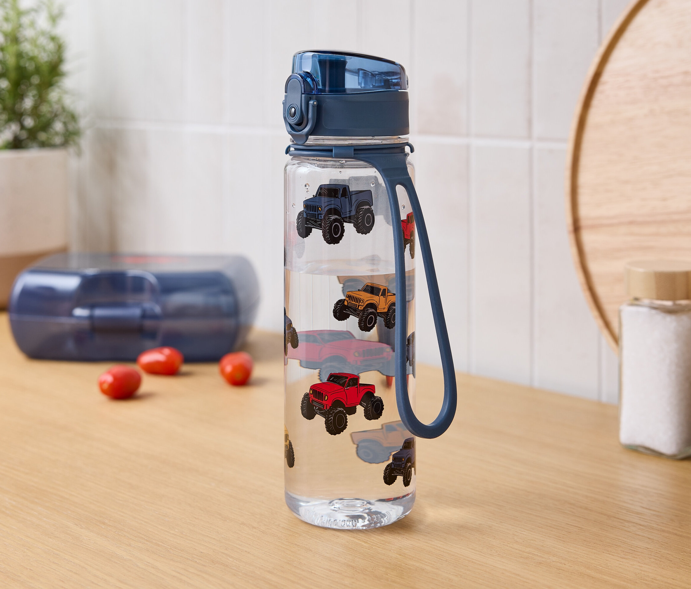 Na stole stojí fľaša s vodou s potlačou autíčok, vedľa nej je krabička na desiatu a koreničky.