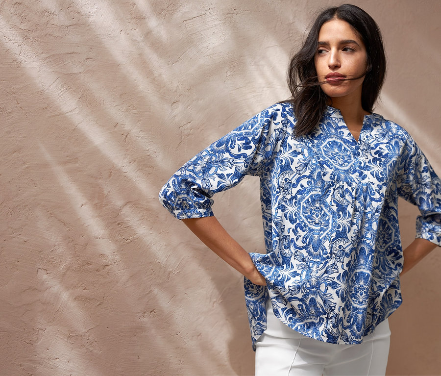 Žena pózuje v tunikovej blúzke s potlačou, bielych sedemosminových elastických nohaviciach a plátenných teniskách. Má tašku na rameno.