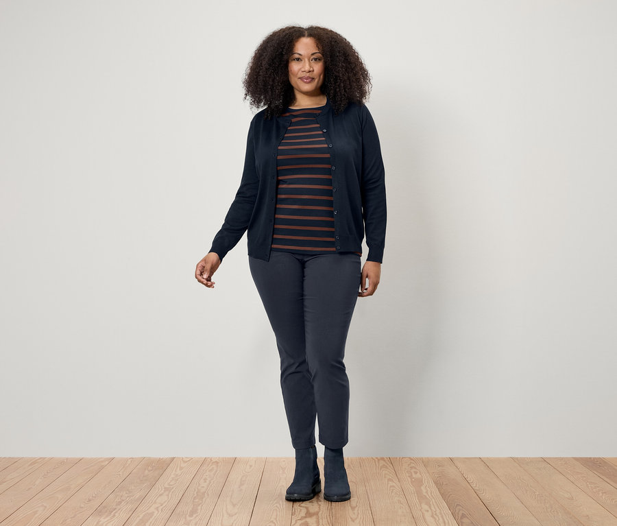 Žena s tmavými kučeravými vlasmi pózuje na drevenej podlahe a má na sebe tmavomodrý kardigán, pruhované tričko a tmavomodré nohavice a topánky.