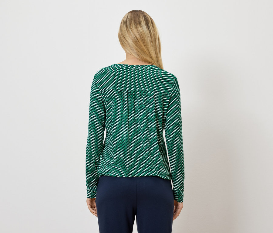 Žena má na sebe zelený pásikavý top s dlhým rukávom a tmavomodré nohavice.