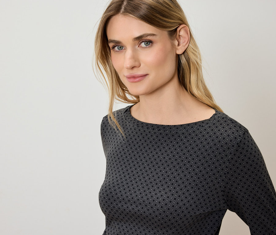 Portrét ženy s blond vlasmi a modrými očami v sivo-čiernom tope s geometrickým vzorom.