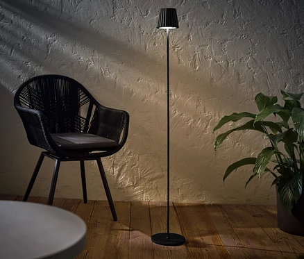 Čierna LED akumulátorová stojaca lampa do exteriéru stojí vedľa čierneho prúteného kresla a črepníkovej rastliny.
