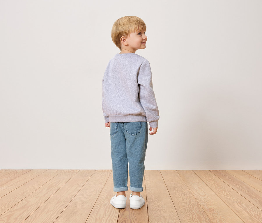 Malý chlapec s blond vlasmi, videný zozadu, má na sebe sivý sveter, modré džínsy a biele topánky.