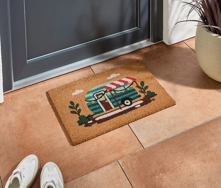 Rohožka s motívom karavanu leží pred sivými dverami na terakotových dlaždiciach.