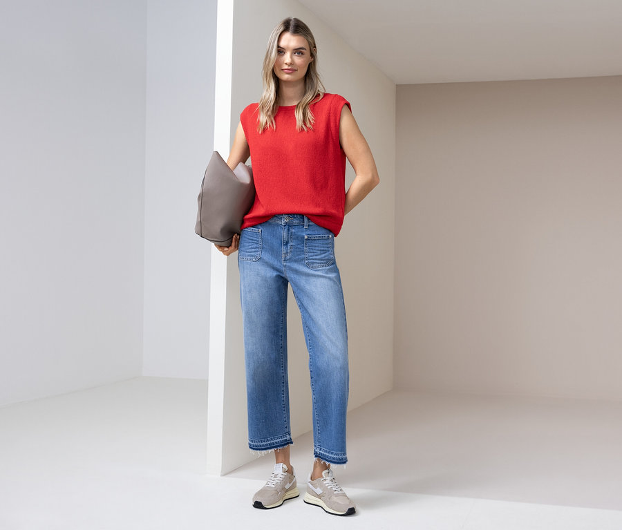 Žena v červenom tope, džínsoch a teniskách stojí so sivou taškou v svetlej miestnosti.