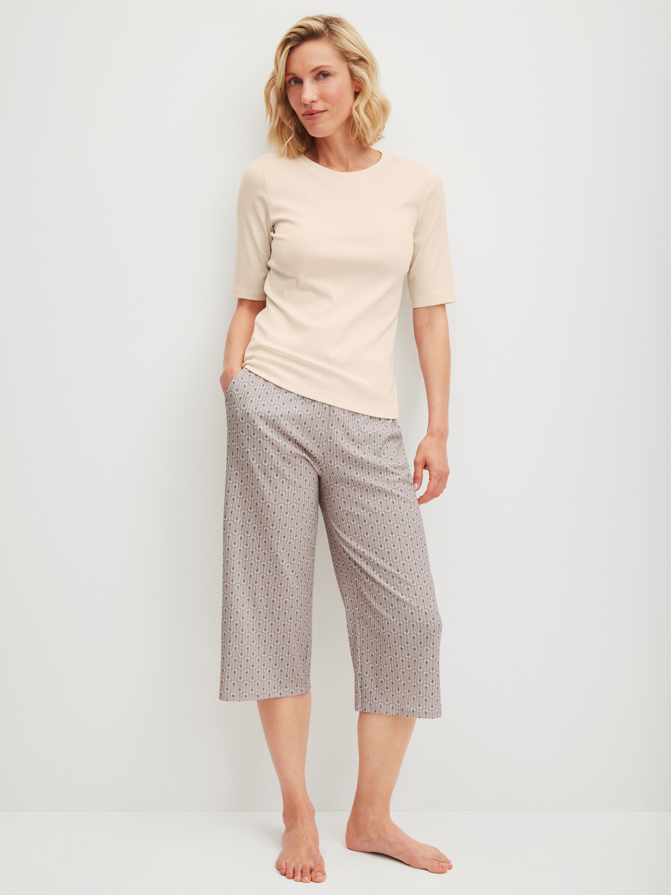 Žena stojí naboso, má na sebe svetlé tričko a vzorované trojštvrťové nohavice.