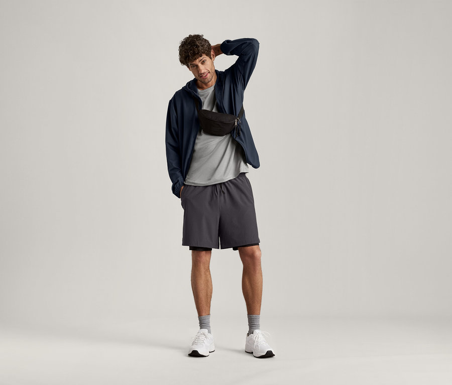 Muž pózuje s rukou na hlave, má na sebe svetlosivé melírované športové tričko, tmavú bundu, šortky a bedrovú tašku.