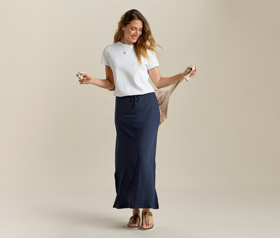 Žena drží béžový kardigán, má na sebe biele tričko a dlhú džersejovú sukňu v námorníckej modrej farbe.