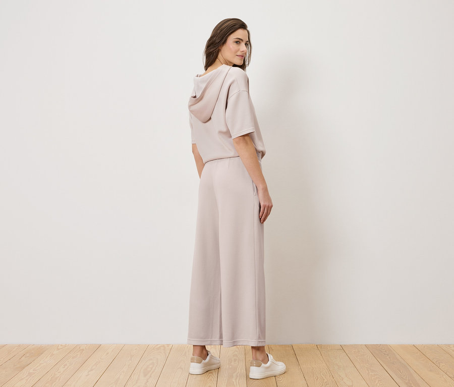 Žena stojí v svetloružovom nohavicovom kostýme a bielych teniskách, otáča hlavu cez rameno.