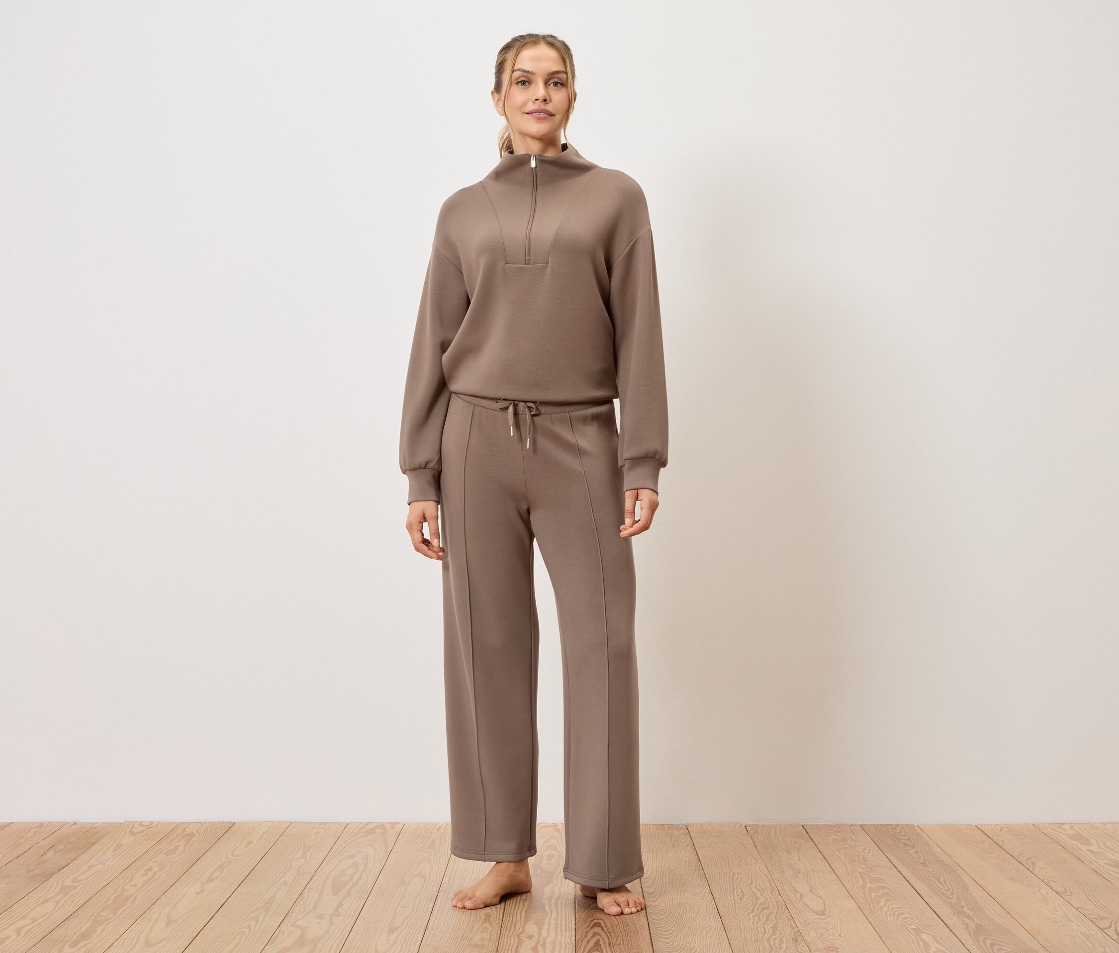 Žena stojí naboso na drevenej podlahe a má na sebe hnedú teplákovú súpravu.