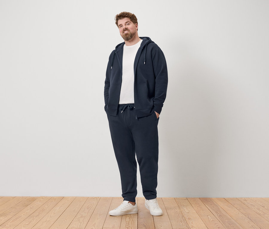 Muž stojí v tmavomodrej teplákovej súprave a bielych teniskách.