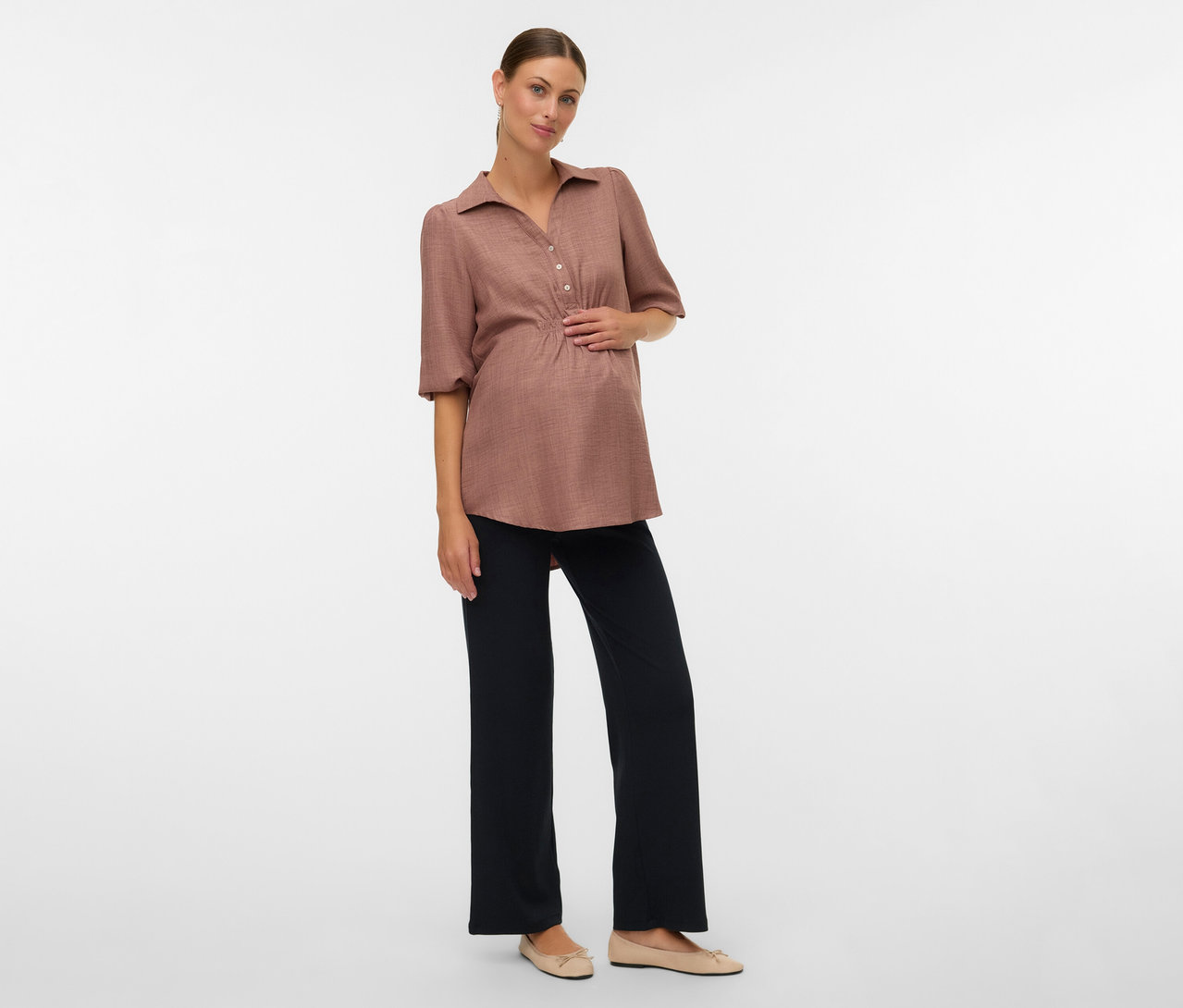 Tehotná žena s hnedou blúzkou a voľnými legínami MAMALICIOUS »MLEMILY« pózuje na bielom pozadí.