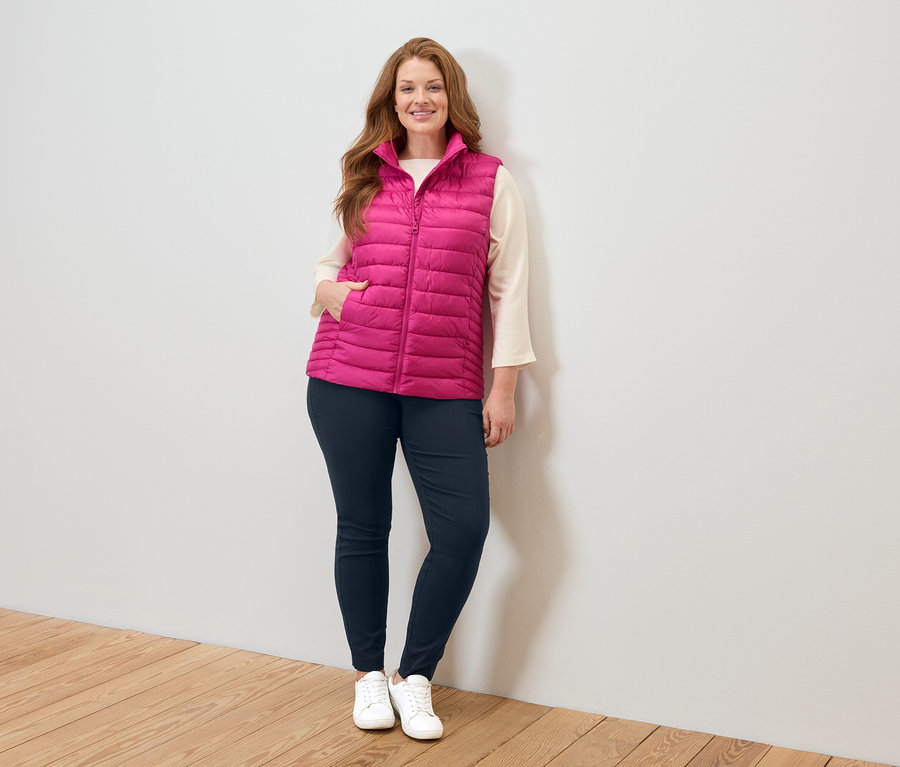 Žena v ružovej prešívanej veste, béžovej blúzke, modrých džínsoch a bielych teniskách stojí pri bielej stene.