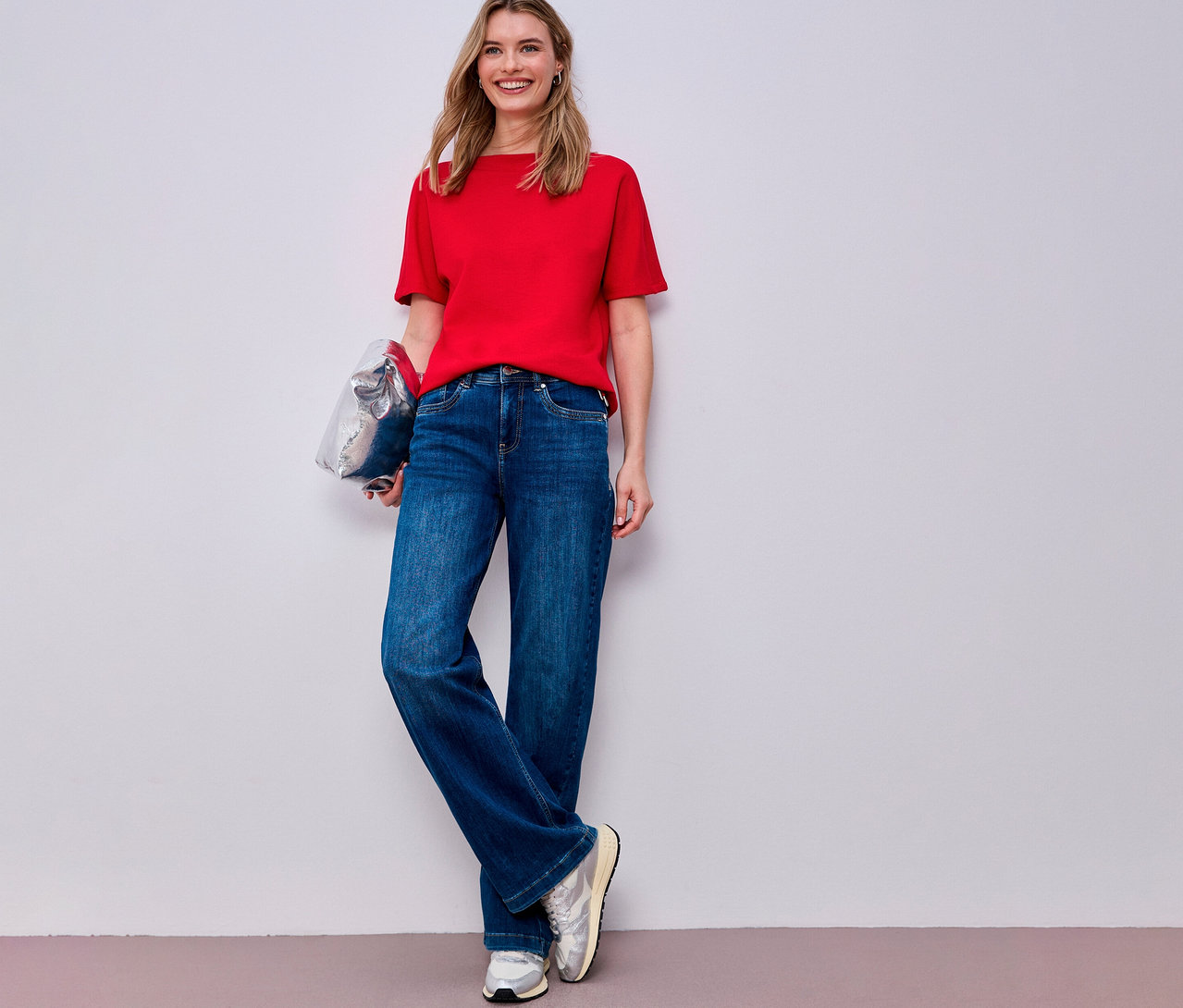 Žena v červenom tričku s krátkym rukávom Street One, modrých džínsoch a strieborných teniskách drží striebornú tašku.