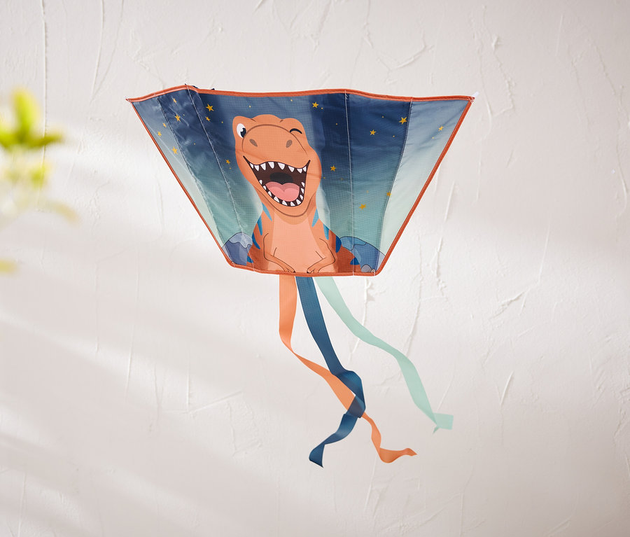 Trapézový šarkan s usmievavým oranžovým dinosaurom a hviezdami visí na textúrovanej bielej stene. Zospodu visia oranžové, tmavomodré a svetlomodré stuhy.