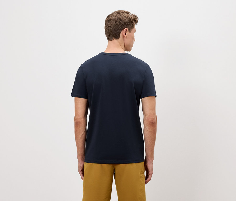 Muž zozadu v námorníckom tričku a zlatých šortkách na jednoduchom pozadí.