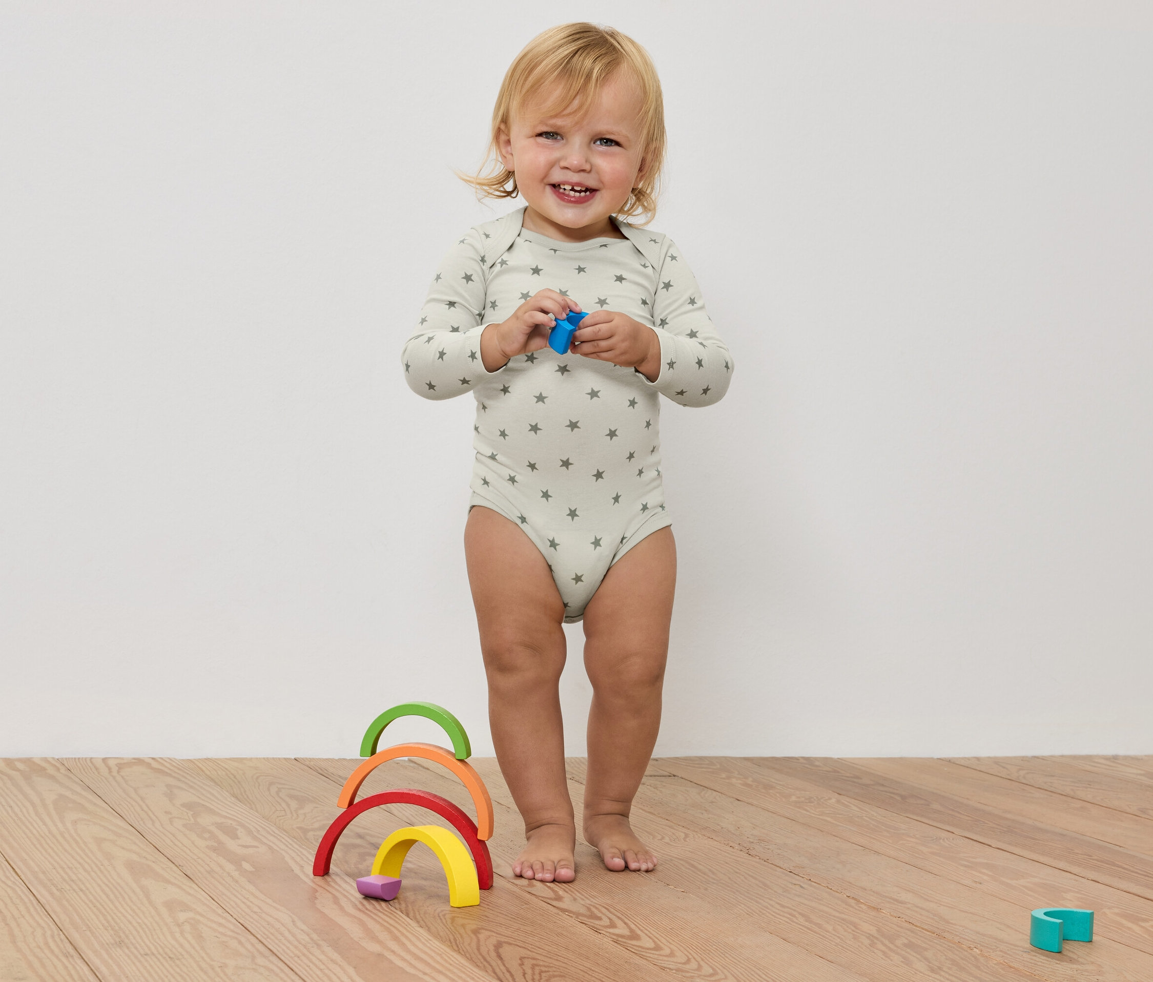 Usmievajúce sa batoľa v body s hviezdami drží hračku v rukách, pred farebnou drevenou dúhou na drevenej podlahe.